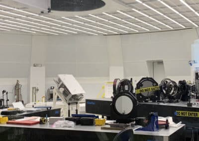 Inside the DKIST Coude Lab is a white, round room filled with specialized benches holding optical elements such as mirrors and lenses. The ceiling has a large hole in the ceiling is where the light passes from the telescope level above into this instrument lab.