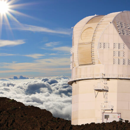 DKIST telescope in in the right foreground, on the red-brown rubbly ground of Haleakalā. In the background, the clouds are clearly below the telescope, and a clear sun is visible in the left background.