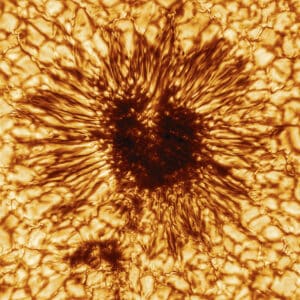 The dark, mottled center of a heart-shaped sunspot is surrounded by the streaking, spidery penumbra and the blotchy, cellular granules of the solar surface. The image is colored shades of orange and brow that enhance the images dimensional feel