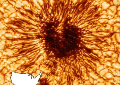 The dark, mottled center of a heart-shaped sunspot is surrounded by the streaking, spidery penumbra and the blotchy, cellular granules of the solar surface. The image is colored shades of orange and brow that enhance the images dimensional feel. The outline of the continental United States sits in the bottom left corner, occupying about the same size as the sunspot.