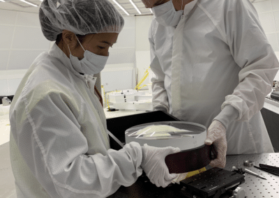 Two scientists in cleanroom gear inspect a lens together.