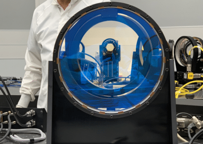 scientists in cleanroom gear stands behind optical lenses that appear blue.