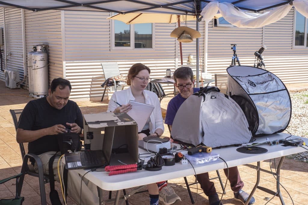 Left to right: Sanjay Gosain, Sarah Bruce, and Kevin Reardon prepare their connections between telescopes, components, and computers in the days leading up to the total solar eclipse. The hoods over the computers help the scientists see their monitors in the harsh morning light. Credit: John Williams/NSO/AURA/NSF